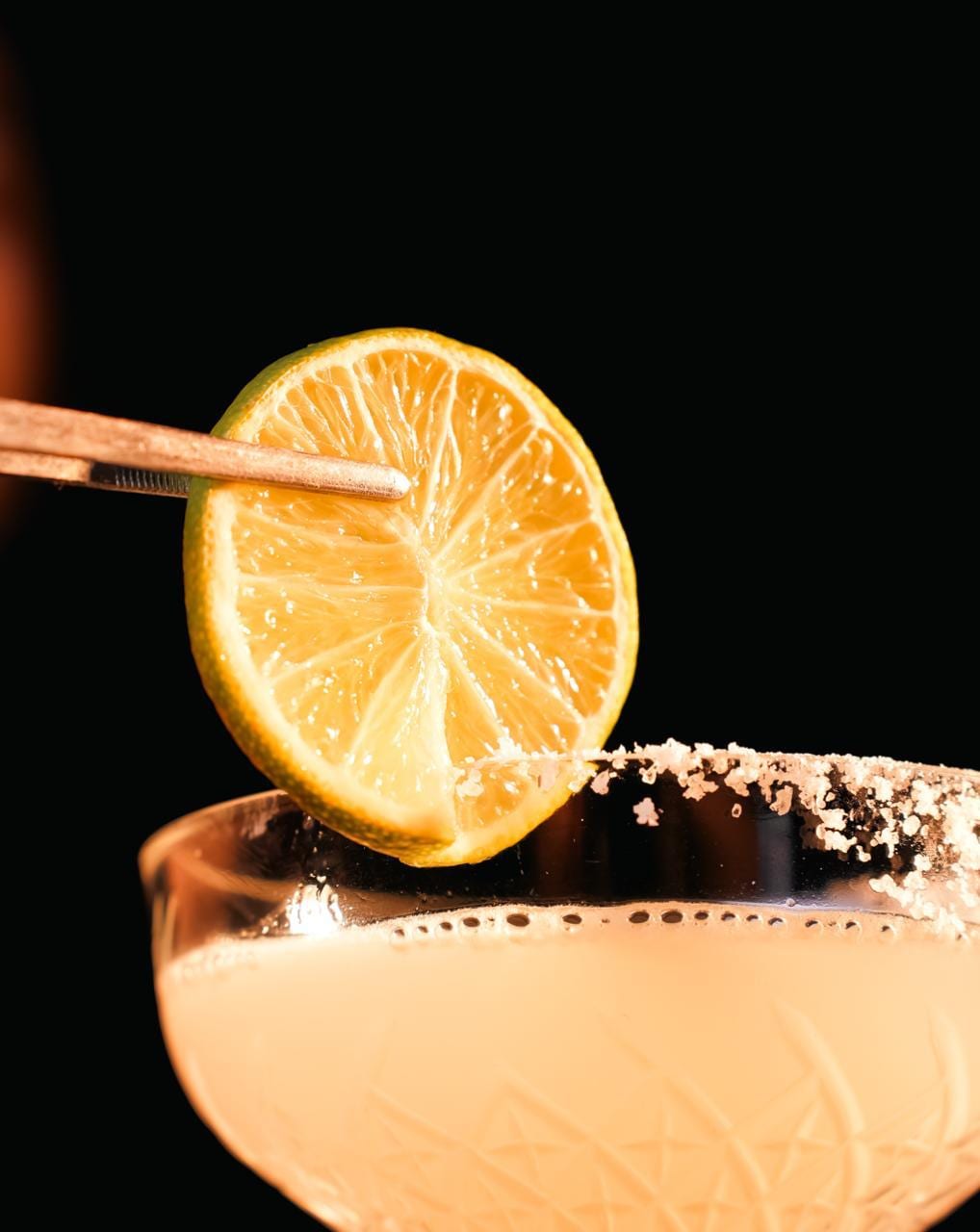 Siyah fonda margarita kadehinde tuz kenarlı kokteyl, maşayla tutulan lime dilimi garnitürü